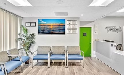 A bright waiting room with blue chairs, a sunset photo on the wall, and a green door